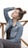 A person wearing a denim jacket, dark top, and dark jeans sits with eyes closed and a relaxed expression. The person has their hair in a bun and is touching their hair with one hand. They are positioned in front of a simple wooden railing against a light background.