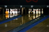 A dimly lit bowling alley with two lanes is depicted, featuring sets of bowling pins arranged neatly at the end of each lane. An orange bowling ball rests on the left lane, while a brown bowling ball is visible on the right. The wooden lanes reflect the pins and balls slightly, adding depth to the scene.