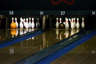 A dimly lit bowling alley with two lanes is depicted, featuring sets of bowling pins arranged neatly at the end of each lane. An orange bowling ball rests on the left lane, while a brown bowling ball is visible on the right. The wooden lanes reflect the pins and balls slightly, adding depth to the scene.