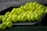 A pile of bright yellow golf balls is lying on an inclined black tray, with some of them scattered on artificial green grass. The golf balls have some black markings and the word 'PRACTICE' written on them. There is a blurry background that includes part of a net and some text.