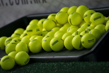 A pile of bright yellow golf balls is lying on an inclined black tray, with some of them scattered on artificial green grass. The golf balls have some black markings and the word 'PRACTICE' written on them. There is a blurry background that includes part of a net and some text.