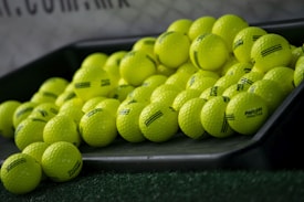 A pile of bright yellow golf balls is lying on an inclined black tray, with some of them scattered on artificial green grass. The golf balls have some black markings and the word 'PRACTICE' written on them. There is a blurry background that includes part of a net and some text.