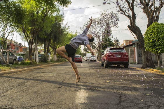 A person is gracefully performing a dance or jumping pose in the middle of a tree-lined street. The individual appears to be caught in mid-air, emphasizing movement and expression. Cars are parked along the sides of the street, and the surrounding trees have dense green foliage.