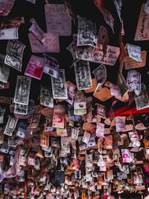 A ceiling covered with various currencies from around the world, each marked with different handwritten messages and drawings. The banknotes and notes create a collage of colors and text, hanging in a busy, cluttered manner.