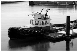 A black and white image of a small tugboat docked at a harbor. The tugboat has a robust design with round fenders attached along the side. It is tied to a wooden dock, and the water surrounding the area is calm. In the background, there are hints of industrial structures.
