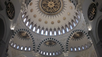 The image features a beautifully ornate ceiling with intricate Islamic calligraphy and geometric patterns. The central dome is adorned with elaborate, symmetrical decorations surrounded by smaller domes, all featuring similar artistic motifs. Light filters through small, evenly spaced windows, creating a serene atmosphere.