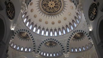 The image features a beautifully ornate ceiling with intricate Islamic calligraphy and geometric patterns. The central dome is adorned with elaborate, symmetrical decorations surrounded by smaller domes, all featuring similar artistic motifs. Light filters through small, evenly spaced windows, creating a serene atmosphere.