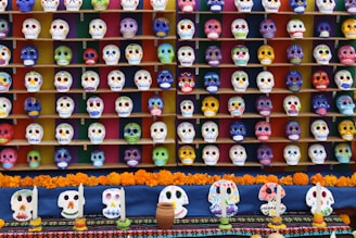 An extensive collection of colorful decorated skulls arranged in multiple rows on shelves, typically used in celebrations for Dia de los Muertos. The skulls are adorned with vibrant designs and patterns, each uniquely painted. In front of the display, there are marigold flowers and candles, adding to the festive atmosphere.