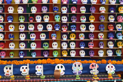 An extensive collection of colorful decorated skulls arranged in multiple rows on shelves, typically used in celebrations for Dia de los Muertos. The skulls are adorned with vibrant designs and patterns, each uniquely painted. In front of the display, there are marigold flowers and candles, adding to the festive atmosphere.