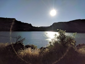 A serene lake scene during sunset or sunrise, with the sun low in the sky casting reflections on the water. Silhouettes of hills and sparse vegetation are visible along the shoreline, adding depth to the tranquil landscape.