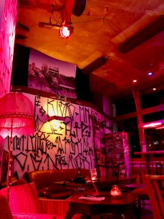 A cozy, dimly lit room with eclectic decor, featuring a mix of vintage furniture and graffiti-covered walls. The space is illuminated by warm red and orange lighting, creating a relaxed and artsy atmosphere. A sofa and seating area are furnished with comfortable chairs, while various art pieces and photographs adorn the walls.