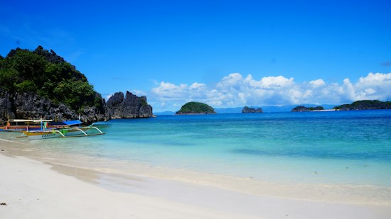 A serene beach scene with calm, clear turquoise water gently lapping onto a sandy shore. Small green islands and dark, rocky outcrops are visible in the distance under a bright blue sky scattered with white clouds. A traditional wooden boat with colorful accents is docked on the shore.