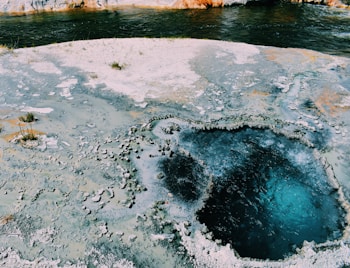 A geothermal hot spring features a deep blue pool surrounded by mineral deposits. The landscape is a mix of rough, textured surfaces and smooth water. Sparse grass and mineral crusts are visible, while a nearby river adds contrast with its darker water.