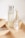 A minimalist arrangement of skincare products on a light wooden tray, featuring a tall frosted bottle with a silver cap, an elegant perfume bottle with a golden neck and glass top, a small opaque bottle, and a gold tube, likely a lipstick.