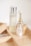 A minimalist arrangement of skincare products on a light wooden tray, featuring a tall frosted bottle with a silver cap, an elegant perfume bottle with a golden neck and glass top, a small opaque bottle, and a gold tube, likely a lipstick.