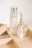 A minimalist arrangement of skincare products on a light wooden tray, featuring a tall frosted bottle with a silver cap, an elegant perfume bottle with a golden neck and glass top, a small opaque bottle, and a gold tube, likely a lipstick.