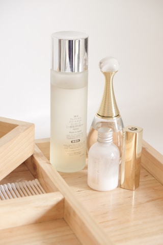 A minimalist arrangement of skincare products on a light wooden tray, featuring a tall frosted bottle with a silver cap, an elegant perfume bottle with a golden neck and glass top, a small opaque bottle, and a gold tube, likely a lipstick.