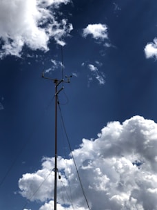 A tall metal weather station tower is set against a backdrop of a deep blue sky. White fluffy clouds drift around, creating a serene and spacious atmosphere. The tower is equipped with instruments, such as anemometers and wind vanes, bound by sturdy cables.