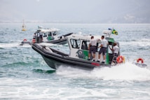 A group of police boats navigating through the sea, with uniformed officers on board. The boats are marked with 'Policia Ambiental', and one of them has a Brazilian flag attached. The water is choppy, creating waves around the boats.