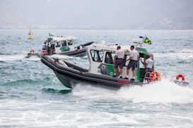 A group of police boats navigating through the sea, with uniformed officers on board. The boats are marked with 'Policia Ambiental', and one of them has a Brazilian flag attached. The water is choppy, creating waves around the boats.