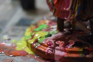 Thick layers of various colorful paints are swirling and pooling together on a surface, creating an abstract and vibrant mixture. The colors blend in a chaotic yet mesmerizing pattern, with glossy and wet textures reflecting light.