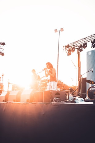 Two musicians perform on an outdoor stage during sunset, one playing the guitar and the other singing into a microphone. The scene is backlit, creating a dramatic silhouette effect, and various stage equipment is visible including speakers and lighting fixtures.