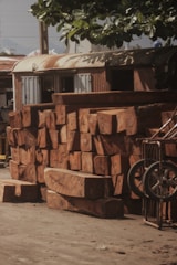 A stack of large, roughly cut wooden logs rests against the side of an old, weathered building with corrugated metal siding. Above, green leaves from a nearby tree provide partial shade. A wheelbarrow with metal wheels is visible on the right side of the image.