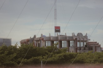 A modern building with a unique architectural design featuring curved and pointed elements on the rooftop, situated behind lush green trees. A red and black sign is mounted on a tall structure above the building.