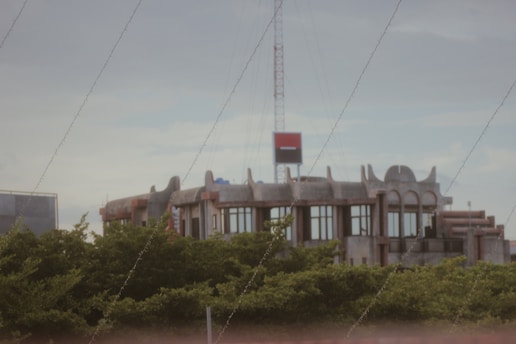 A modern building with a unique architectural design featuring curved and pointed elements on the rooftop, situated behind lush green trees. A red and black sign is mounted on a tall structure above the building.