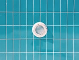 A circular pool light is embedded in a wall of blue rectangular tiles with thin white grout lines.