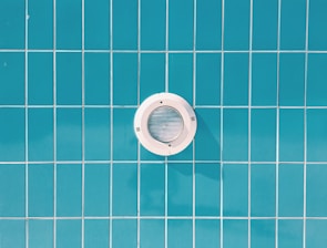 A circular pool light is embedded in a wall of blue rectangular tiles with thin white grout lines.