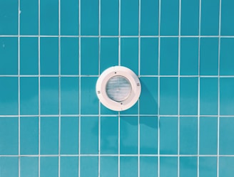 A circular pool light is embedded in a wall of blue rectangular tiles with thin white grout lines.