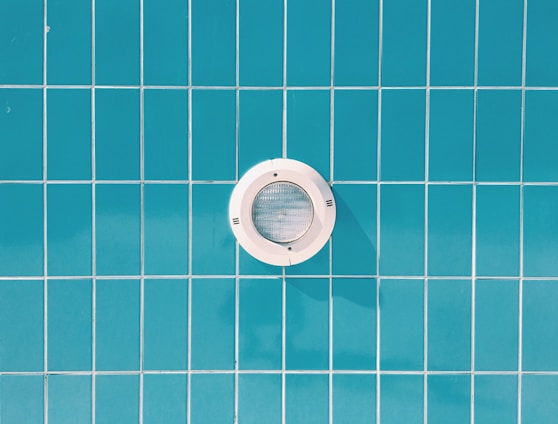 A circular pool light is embedded in a wall of blue rectangular tiles with thin white grout lines.