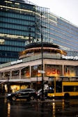 An urban street scene featuring a modern glass building with illuminated signs. The ground level hosts a Superdry store with prominent signage. A double-decker bus and several cars are present on the wet street in front.