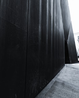 Black metal panels forming an angular and modern architectural structure with a passageway to a lighter area visible at the far end. The surface of the panels is textured and scratched, adding a worn aspect. A partial view of a building and trees can be seen in the background.