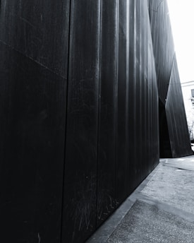 Black metal panels forming an angular and modern architectural structure with a passageway to a lighter area visible at the far end. The surface of the panels is textured and scratched, adding a worn aspect. A partial view of a building and trees can be seen in the background.