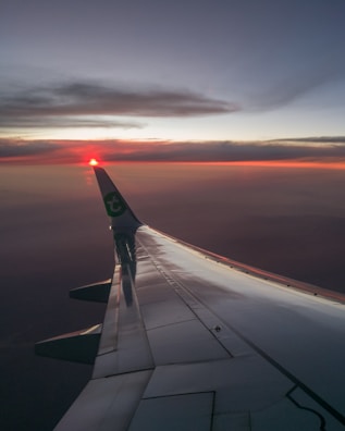 An airplane wing is visible in the foreground, with a sunset or sunrise creating a warm glow on the horizon. The sky transitions from dark blues to lighter shades near the horizon, with clouds reflecting the reddish hues from the sun. The airplane's wingtip has a logo with a green color.