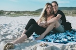 A couple sits together on a patterned blanket spread over sandy beach with their legs intertwined, smiling at the camera. The beach has a backdrop of grassy dunes under a blue sky.