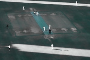 Aerial view of a cricket match in progress on a pitch. Several players in white uniforms are positioned around the field, with one player bowling towards the batsman. The pitch and surrounding field have a slightly worn appearance.