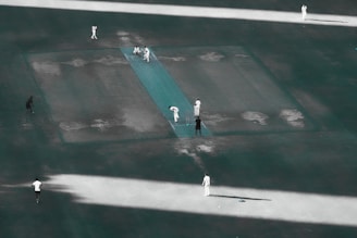 Aerial view of a cricket match in progress on a pitch. Several players in white uniforms are positioned around the field, with one player bowling towards the batsman. The pitch and surrounding field have a slightly worn appearance.