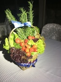 A wicker basket filled with fresh greens such as lettuce and herbs, adorned with ripe red strawberries. The basket is decorated with a blue ribbon and rests on a patterned tablecloth.