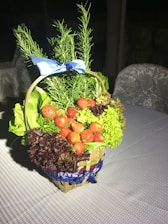 A wicker basket filled with fresh greens such as lettuce and herbs, adorned with ripe red strawberries. The basket is decorated with a blue ribbon and rests on a patterned tablecloth.