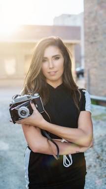 A black-and-white portrait of a photographer holding a vintage camera, standing in a sunlit urban alley.