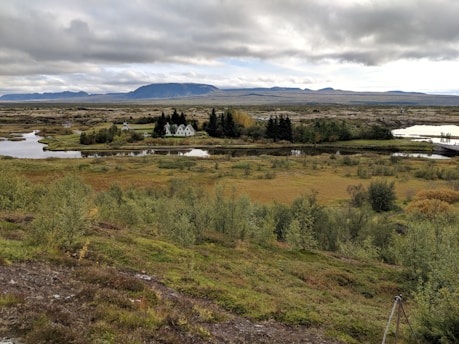 A peaceful rural landscape featuring a small cluster of white houses surrounded by dense trees. In the background, a large mountain range extends beneath a cloudy sky. The foreground is composed of lush greenery and a winding river reflecting the sky.