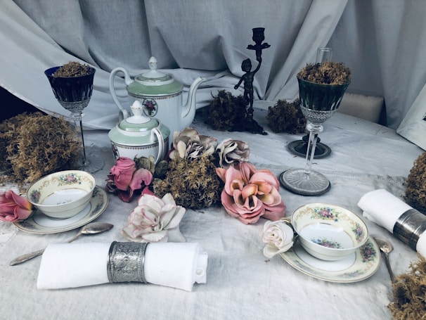 An ornate and vintage-style table setting featuring delicate porcelain teapots and teacups with floral patterns. The arrangement includes cloth napkins with silver holders, moss-filled glasses, and artificial flowers like roses scattered around. A small bronze statuette is placed in the background along with an elegant tablecloth.