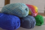 Several skeins of yarn in various colors are neatly arranged. The yarn includes shades of blue, green, and a multicolored skein with pink and yellow hues. The fibers are tightly wound, creating a textured appearance.