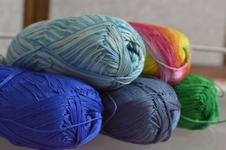 Several skeins of yarn in various colors are neatly arranged. The yarn includes shades of blue, green, and a multicolored skein with pink and yellow hues. The fibers are tightly wound, creating a textured appearance.
