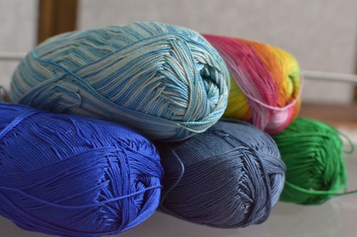 Several skeins of yarn in various colors are neatly arranged. The yarn includes shades of blue, green, and a multicolored skein with pink and yellow hues. The fibers are tightly wound, creating a textured appearance.