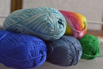 Several skeins of yarn in various colors are neatly arranged. The yarn includes shades of blue, green, and a multicolored skein with pink and yellow hues. The fibers are tightly wound, creating a textured appearance.