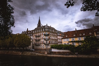 A dramatic scene featuring historic European architecture alongside a river, with overcast skies creating a moody atmosphere. Leafy trees frame the scene from above and to the side.